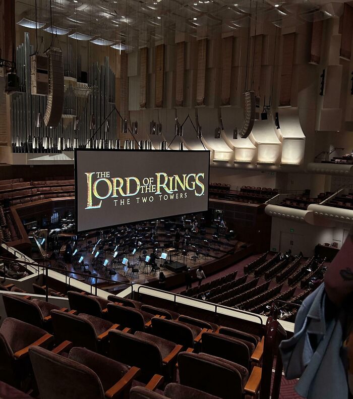 Concert hall setup with orchestra and large screen showing The Lord of the Rings, inspiring big and small dreams realized.