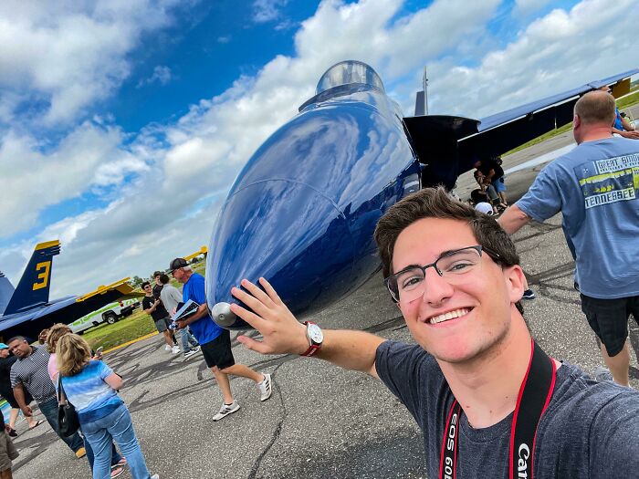 Young man with glasses smiling and posing by a jet aircraft, inspiring dreams turned into reality on the internet.