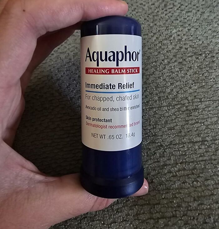 Hand holding Aquaphor healing balm stick for immediate relief on textured gray surface during Black Friday deals week.