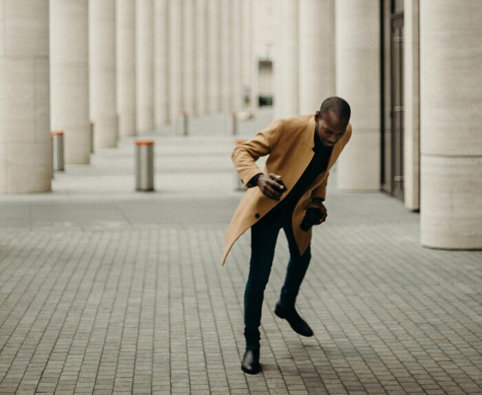 Man in brown coat caught mid-step walking through columns, illustrating relatable but ridiculous moments we’re all probably guilty of.