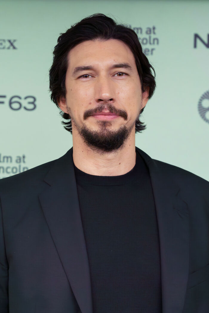 Adam Driver wearing a black shirt and blazer, posing at a film event with career highlights in the background.
