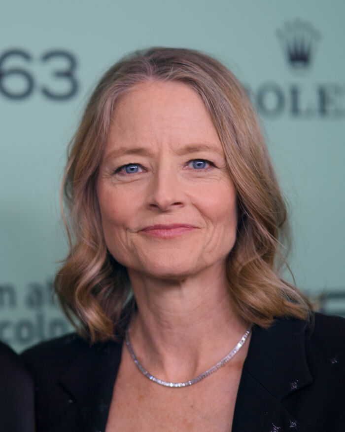 Jodie Foster at a formal event, smiling softly with wavy hair and wearing a black outfit and silver necklace.
