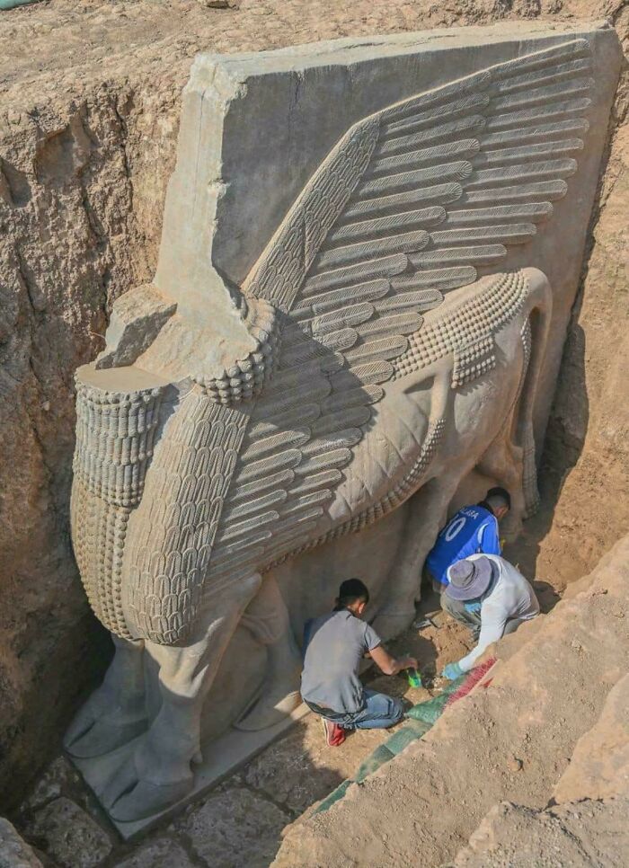 Archaeologists carefully excavating a large ancient winged bull relief at an archeology dig site.