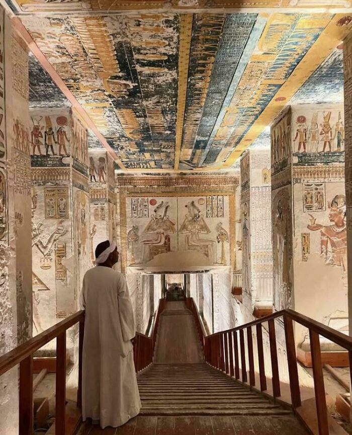 Man in traditional clothing inside an ancient Egyptian tomb with detailed archeology wall paintings and carvings.