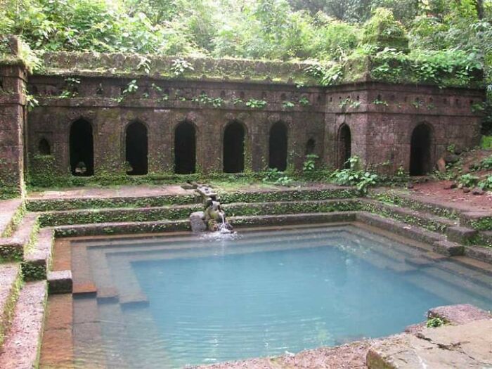 Ancient archeology site with stone structures and a clear blue water pool surrounded by lush greenery and moss.