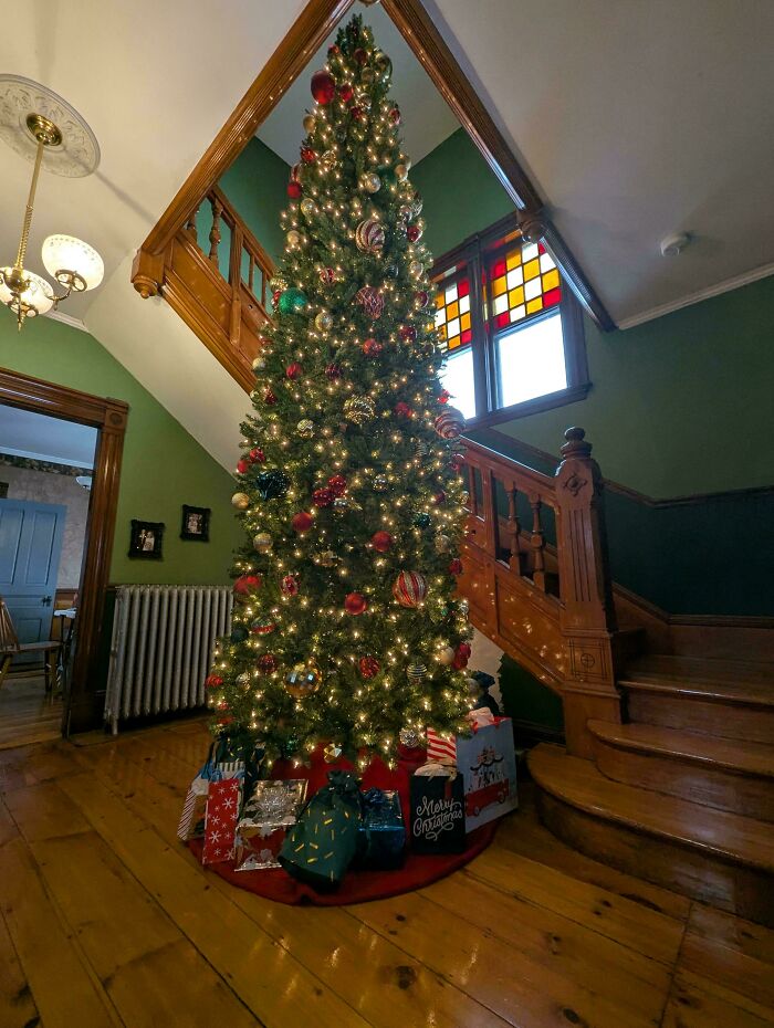Tall decorated Christmas tree with lights and presents in a hallway of a beautiful old house showcasing craftsmanship.