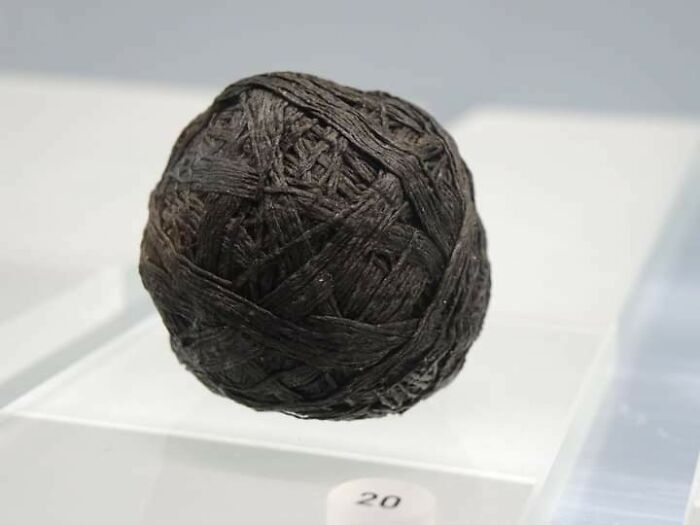 Ancient archaeological artifact resembling a tightly wound fiber ball displayed on a clear stand in a museum exhibit.