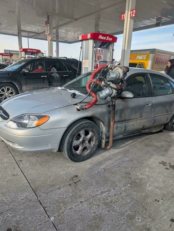 Silver car at gas station with large makeshift engine modification on hood, showing stupidity on wheels attempt to look cooler.