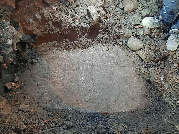 Ancient carved stone slab partially uncovered in an archaeological dig site with workers' feet visible nearby.