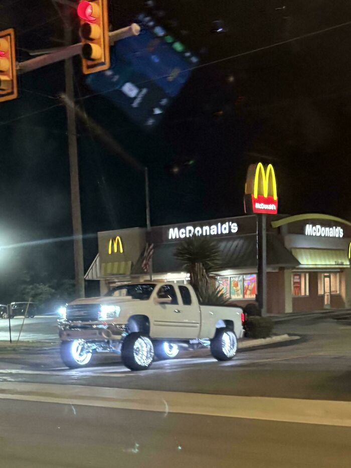 Lifted white pickup truck with glowing, illuminated rims at night in front of a McDonald's, showing stupidity on wheels modification.