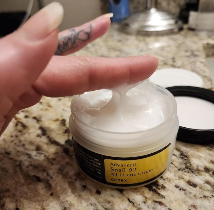 Hand dipping finger into COSRX Advanced Snail 92 cream jar on granite countertop, showcasing beauty restocking product.