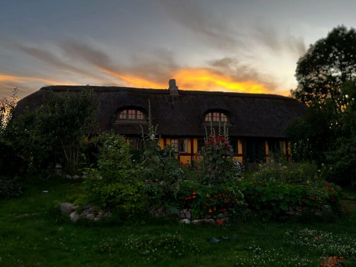 Beautiful old house with thatched roof surrounded by lush garden at sunset, showcasing timeless craftsmanship and design.