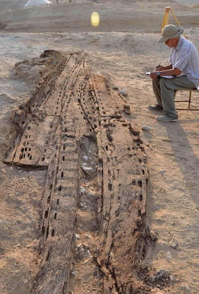 Archeologist sketching detailed ancient wooden shipwreck remains uncovered at an excavation site in a desert region.