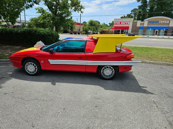 Red car with a large yellow wing and silver side pipes, an example of people trying to make their cars cooler but failing.
