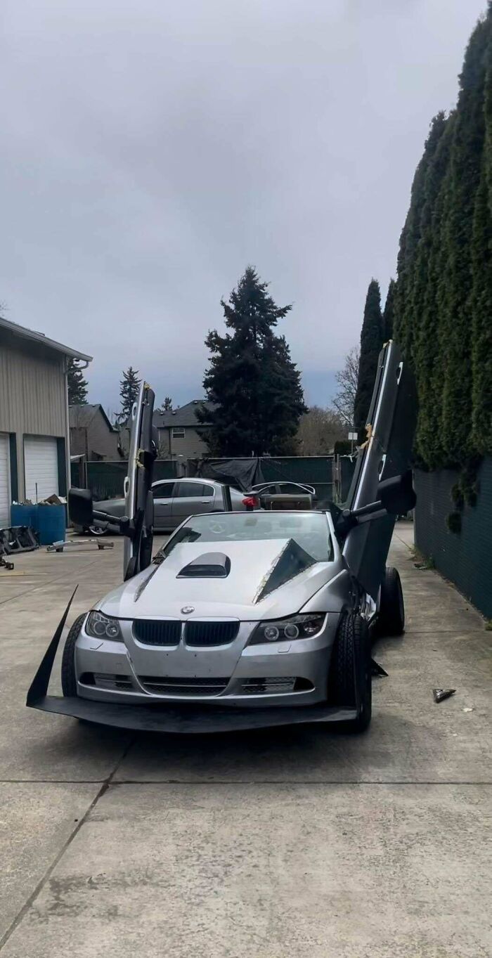 Silver BMW with vertical doors and exaggerated body modifications parked in a driveway, showing failed car coolness attempt.
