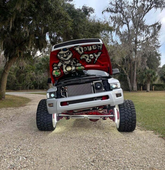Off-road truck with oversized tires and custom graffiti under the hood showing a tough boy design on a rural gravel path.