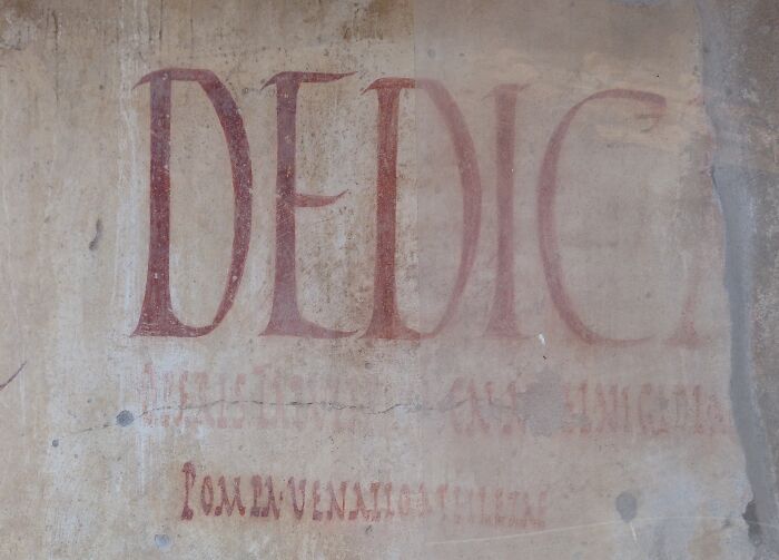 Faded ancient wall inscription with the word DEDIC in red, related to hilarious historical events.