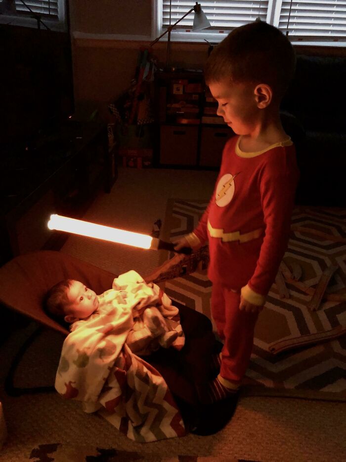 Young boy in red pajamas holding glowing toy sword, standing beside baby sibling wrapped in blanket indoors at night.