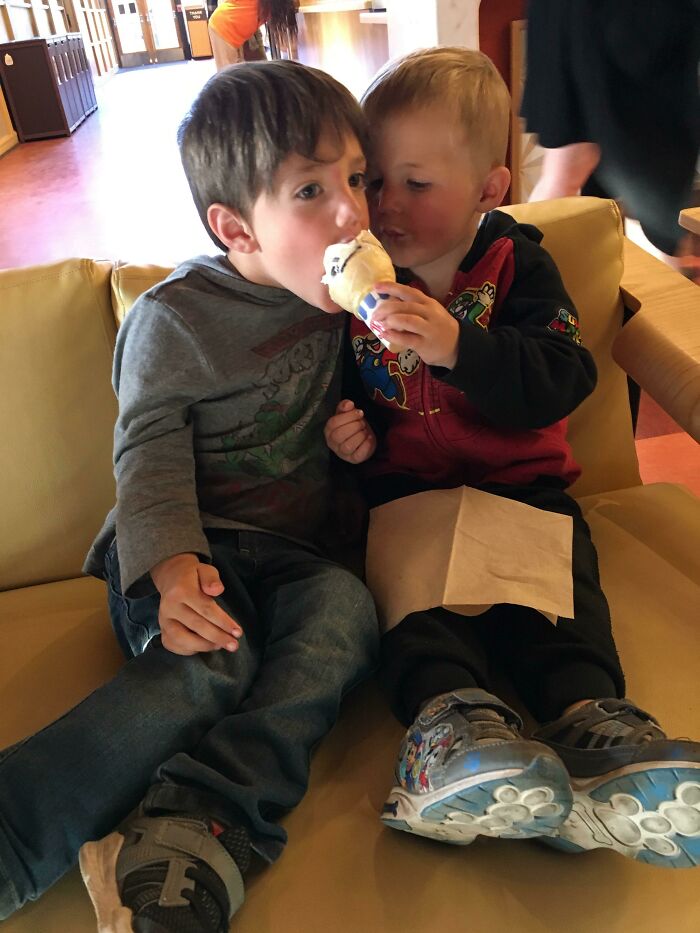 Two young siblings sharing an ice cream cone while sitting closely together on a couch indoors.
