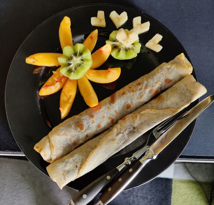 Two rolled crepes on a black plate with kiwi, peach slices, and banana hearts, perfect for a grateful sibling breakfast.