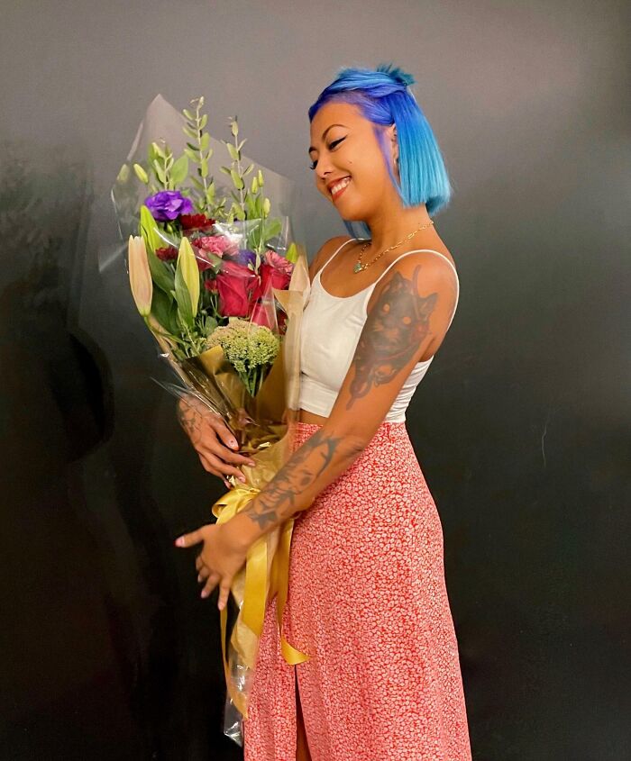 Woman with blue hair and tattoos smiling while holding a bouquet of flowers, inspiring others by turning dreams into reality.