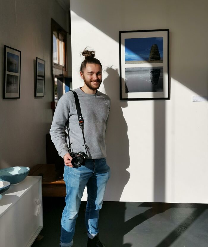 Young man with camera smiling beside framed photos in gallery, showcasing people turning dreams into reality and inspiring others online