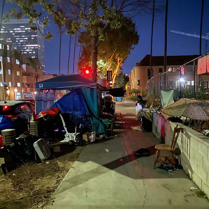Homeless encampment at night on a city sidewalk in Miami with tents, chairs, and nearby buildings under artificial lights.