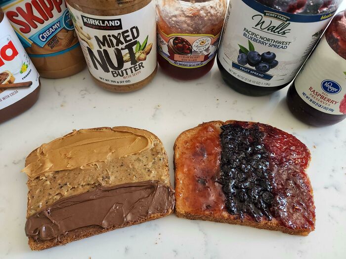 Two slices of bread with peanut butter, mixed nut butter, chocolate spread, and three types of fruit jam on a white countertop.