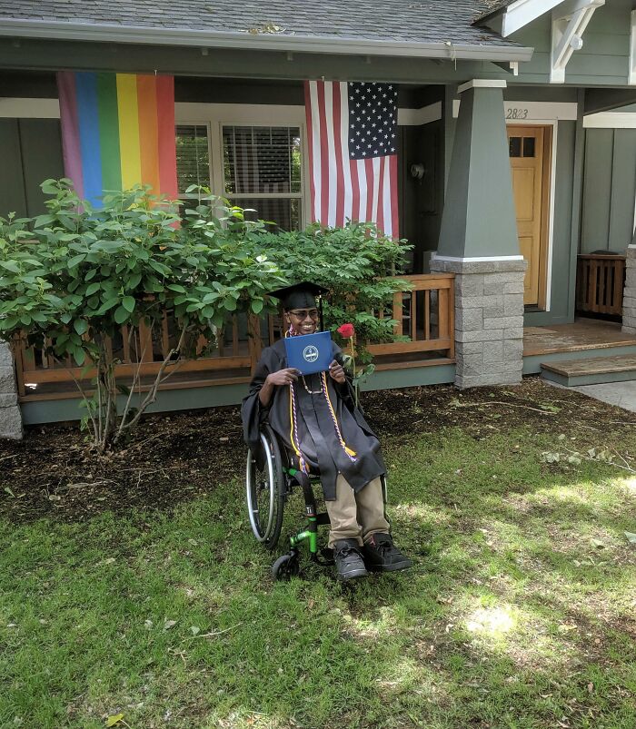 Graduate in cap and gown sitting in wheelchair holding diploma and rose in front of house, inspiring dreams turned into reality.
