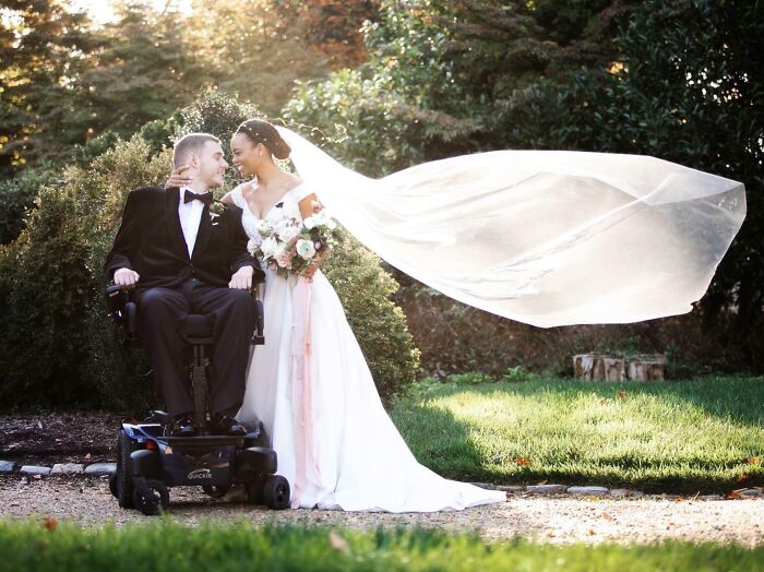 Bride with flowing veil embracing groom in wheelchair outdoors, capturing big and small dreams turned into reality.