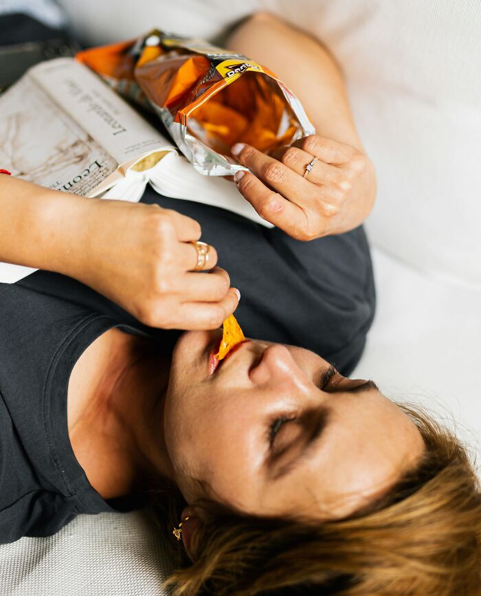 Person lying on couch eating chips while reading a magazine, illustrating relatable guilty habits and everyday moments.