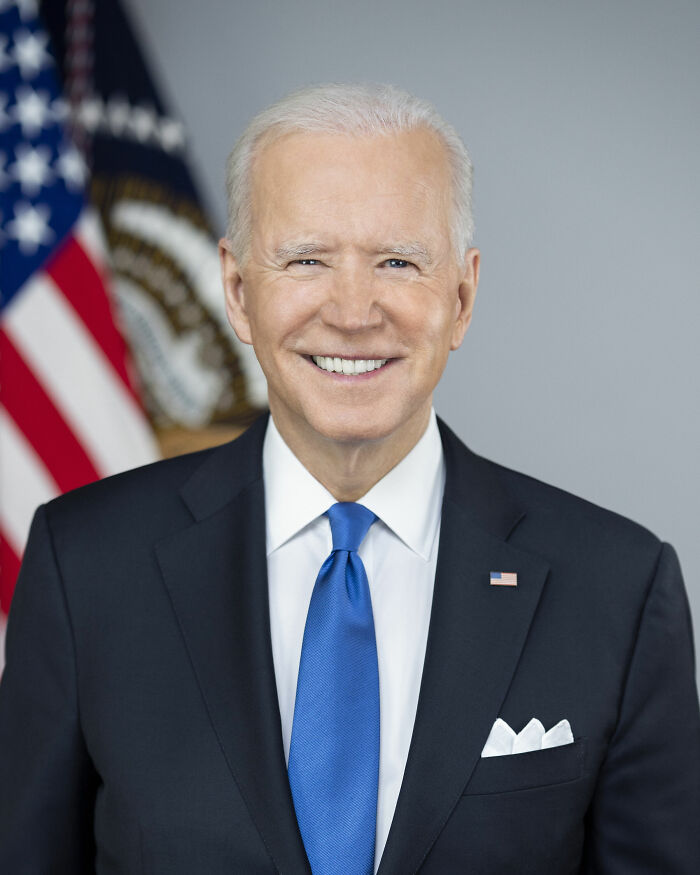 Joe Biden smiling in a dark suit and blue tie with American flag and presidential seal in the background.