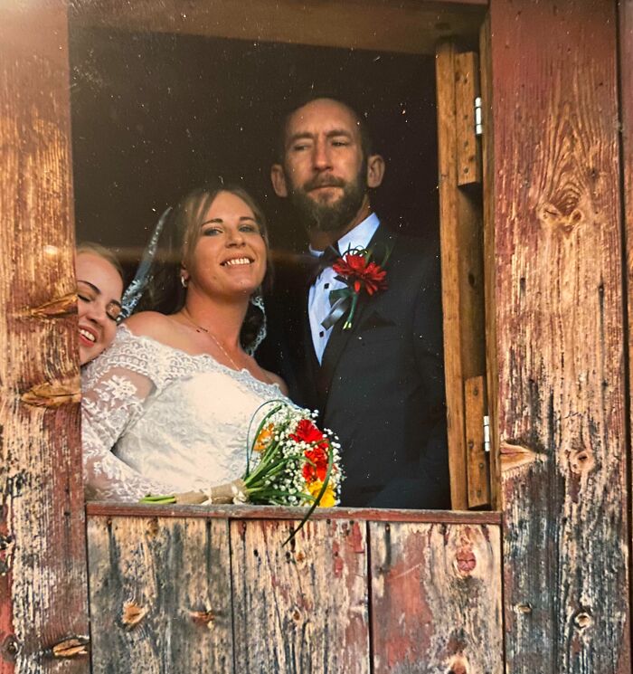 Bride and groom posing in rustic window frame with a bridesmaid photobombing, capturing an epic photobomb moment.