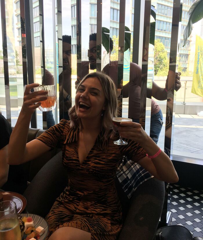 Woman laughing and holding drinks while two men photobomb behind reflective blinds in an epic photobomb moment.