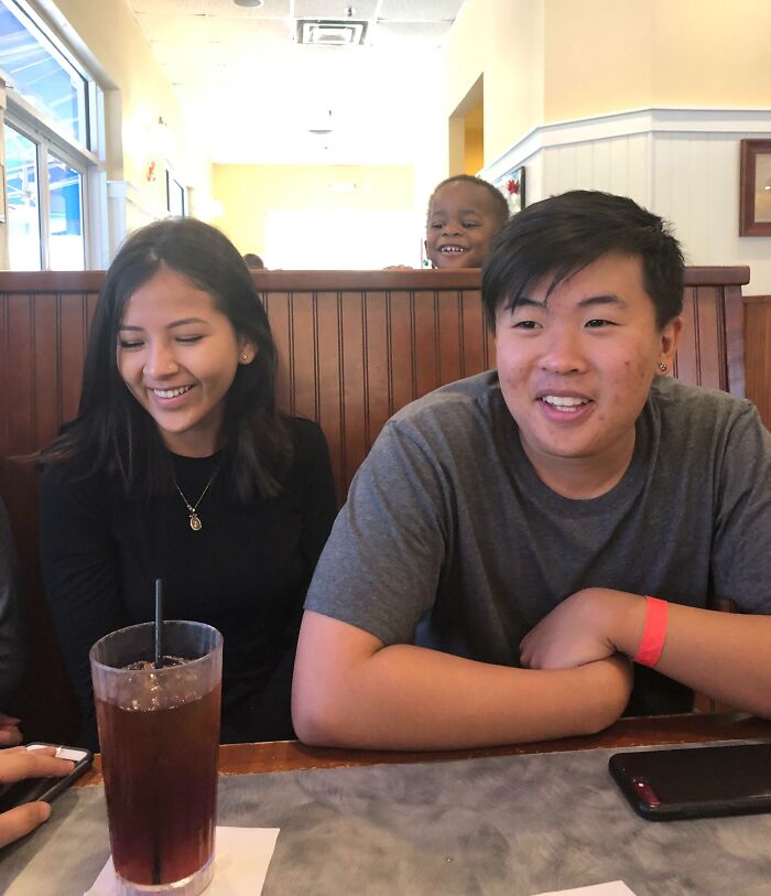 A young boy photobombing two friends sitting at a restaurant table, creating an epic photobomb moment.
