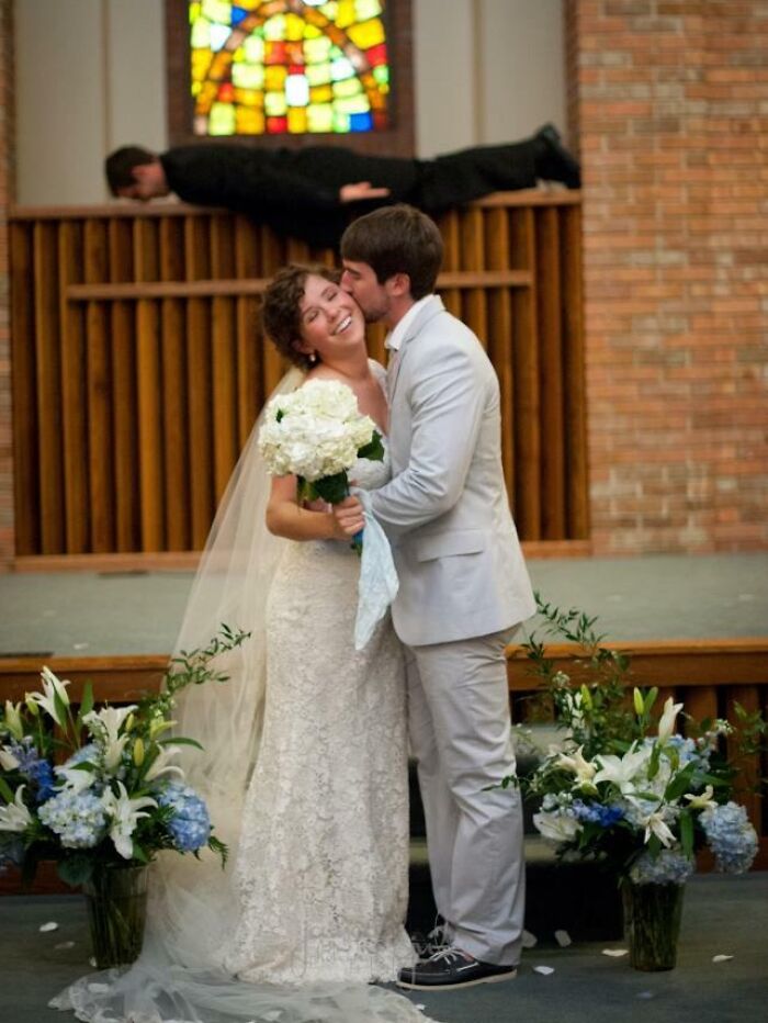 Bride and groom sharing a kiss at wedding while a man photobombs by lying horizontally behind them, capturing epic photobombs.