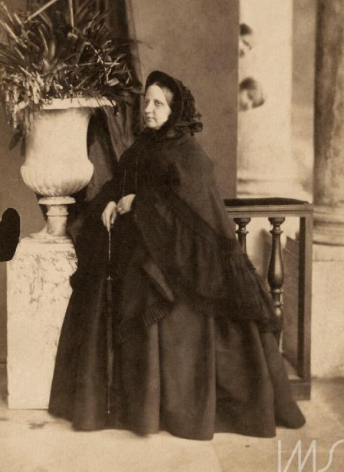 Victorian woman in dark dress and hood posing beside large vase while a child photobombs in the background, epic photobombs.