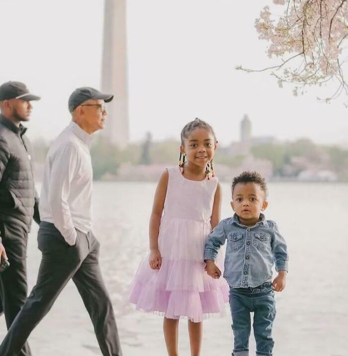 Two young children holding hands near a waterfront while two men walk behind in an epic photobomb moment.