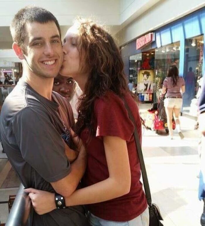 Couple sharing a kiss in a mall with a photobomber making a funny face behind them, capturing an epic photobomb moment.
