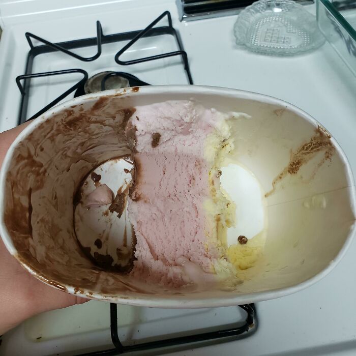 Half-eaten ice cream container held over a stove, showing chocolate, strawberry, and vanilla flavors, playful and wholesome moment.
