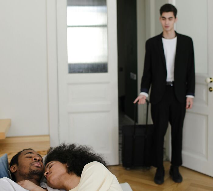 Man in black suit with suitcase looking upset while couple lies together on bed, illustrating brutal revenge on an ex concept.