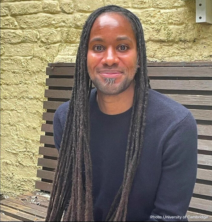 Man with long dreadlocks sitting on a wooden bench against a yellow brick wall, representing wholesome internet moments.