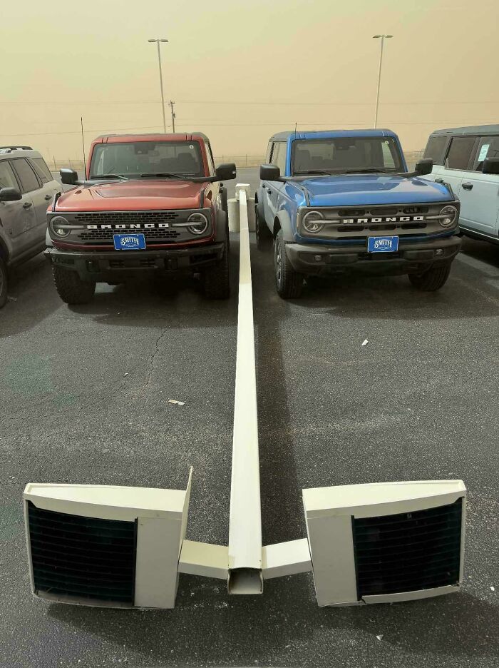Two Ford Broncos perfectly aligned with a parking barrier and two air conditioning units in a lot.