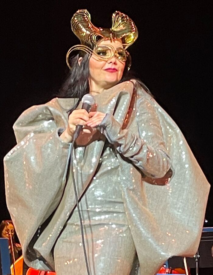 Bj&ouml;rk performing live on stage wearing an elaborate metallic costume and holding a microphone during a concert.