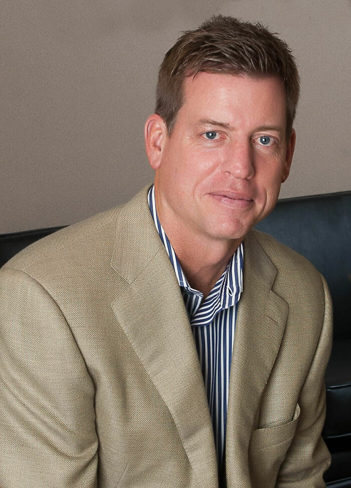 Troy Aikman in a beige blazer and striped shirt, seated indoors, representing his bio and career highlights.