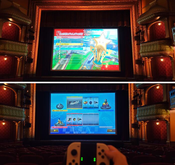 Theater screen showing a video game leaderboard and gameplay, with a player holding controllers, reflecting employee pranks and jokes.