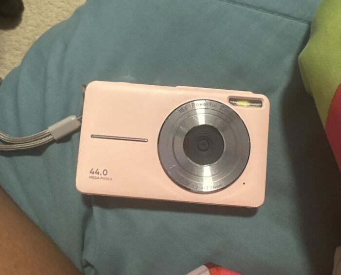 Compact pink digital camera with 44 megapixels placed on a blue fabric surface, featuring tech details and a wrist strap.