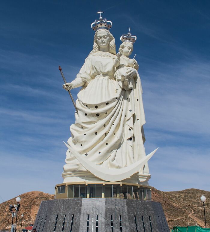 Large white sculpture of a crowned woman holding a child, standing on a stone pedestal against a clear blue sky.