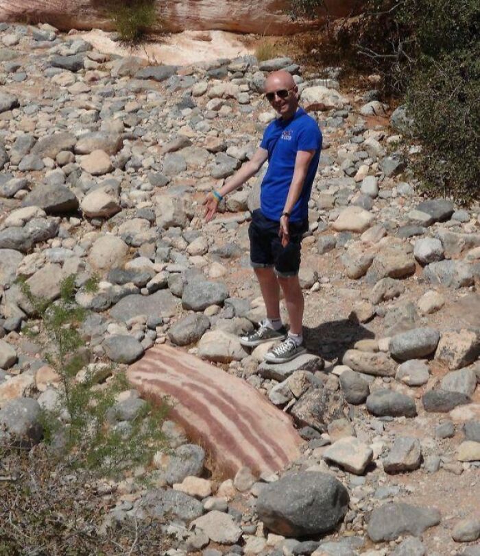 Man in blue shirt standing on rocky ground pointing at a large rock resembling striped bacon in forbidden foods context