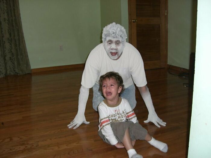 Man with white face paint playfully chasing young boy crying on wooden floor, showcasing wholesome photos of men never growing up.